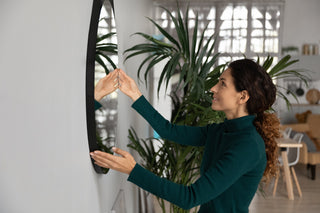 Woman in a green sweater adjusting a round black-framed wall mirror, surrounded by indoor plants in a modern living space.