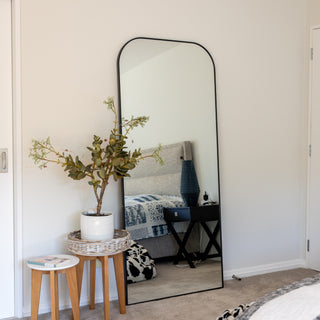The Arcadia Full-Length Mirror 180x80 features a sleek, arched black frame, complementing a contemporary bedroom setting. Placed against a white wall, it enhances the space by reflecting natural light and creating an open, airy feel. A stylish and functional home décor piece. | C&F Creation