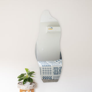 Front view of the Cascade Frameless Full-Length Curvy Mirror in 160 x 60cm, wall-mounted in a bright bedroom. The mirror’s abstract silhouette stands out against a clean white wall, reflecting a bed with patterned blue bedding and part of a window. A potted plant on a woven basket stand adds a fresh, natural touch beneath the mirror.