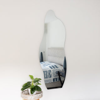 The Cascade Frameless Full-Length Curvy Mirror in 160 x 60cm is wall-mounted in a modern bedroom. Its organic silhouette adds a soft artistic touch, reflecting a neatly styled bed, black bedside table, and ceramic lamp for a calm, minimalist vibe. A lush plant in a woven basket sits below the mirror for a natural accent.