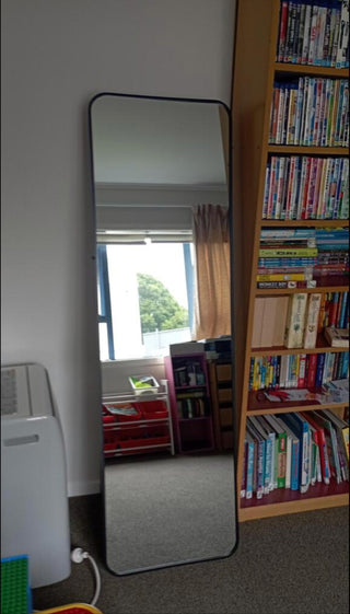 A customer photo of the Lithe Rounded Corner Full-Length Mirror (150x45cm) placed in a room with bookshelves and a window. Accompanied by a 5-star review praising the mirror’s quality and accurate reflection without distortion.