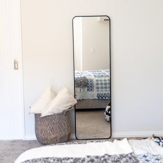 The Lithe X Full-Length Mirror (155x45cm) with a slim black frame, leaning stylishly against a white wall in a modern bedroom. This versatile mirror can be wall-mounted, used as a floor mirror, or stand freely. Complemented by soft décor elements, including a woven basket and cozy pillows. | C&F Creation