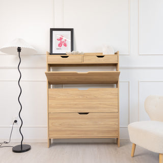 Risa Slim Shoe Cabinet in oak finish shown with one of the three shoe compartments open, highlighting its tilt-down storage function. The cabinet also features two top drawers for smaller items, styled in a bright living space with a floor lamp, artwork, and accent chair.