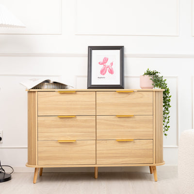 The Ruelle 6-Drawer dresser in warm oak finish, featuring fluted side detailing and minimalist handles, styled with books, artwork, and greenery in a modern interior.