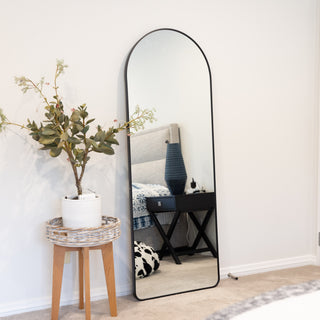 Svelte X Arched Full-Length Mirror with sleek black frame leaning against a bedroom wall, reflecting a cosy modern interior with a nightstand and blue decor. C&F Creation.