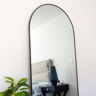 A sleek Titan arched full-length mirror (180x80 cm) mounted on a minimalist white wall. The black frame complements the contemporary bedroom décor, featuring a bedside table with a stylish blue lamp and a lush green plant. | C&F Creation