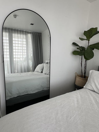 A stunning Titan Arched Full-Length Mirror (180x80cm) elegantly placed in a modern bedroom, reflecting a neatly made bed with soft white bedding and a stylish green indoor plant. This customer-favorite mirror enhances the room’s brightness and space while adding a sleek, minimalist touch. A 5-star review from a satisfied customer praises the high-quality packaging, professional delivery service, and outstanding value.