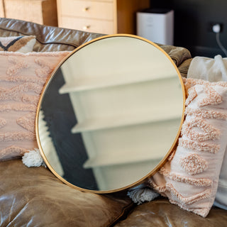 Valor Round Wall Mirror with a luxurious gold aluminium frame, placed on a leather sofa decorated with textured cushions for a warm and elegant living space | C&F Creation