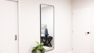 A full-length black-framed mirror securely mounted on a bedroom wall, showing a clean and professional installation with a tidy reflection of a bedside table and decor — ideal for demonstrating wall-mount setup.
