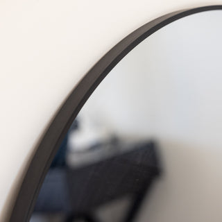 A detailed close-up of the Zia Round Wall Mirror, showcasing its sleek black aluminium alloy frame and flawless reflective surface. The minimalist design blends seamlessly with modern interiors, enhancing both functionality and aesthetics. The background subtly features a stylish bedroom setup with contemporary decor. | C&F Creation