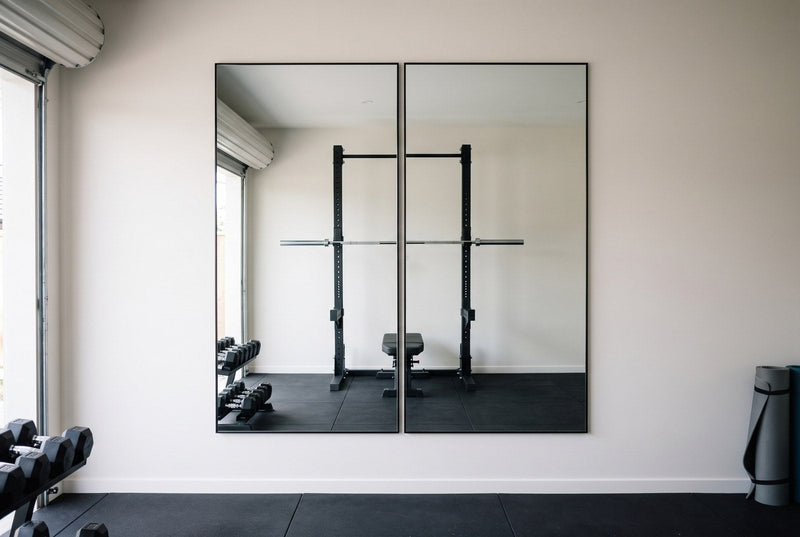Two black framed Aldren X 200 x 100cm mirrors hung upright side by side on a white wall in a New Zealand garage gym, reflecting a squat rack, barbell and bench on black rubber flooring