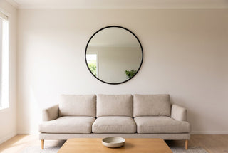 Aure 100cm round mirror with a slim black frame hung centred above an oatmeal linen three seat sofa in a New Zealand living room, with a clear band of wall between the top of the frame and the ceiling