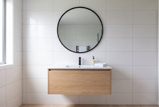 Aure 100cm round mirror with a slim black frame hung above an oak vanity on a New Zealand bathroom wall tiled in 600 by 300mm white tiles with straight grout lines