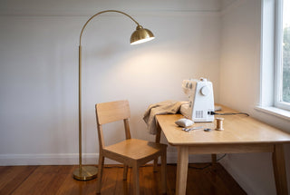 Orsay gold arc floor lamp standing to the left of an empty timber chair at a sewing table in a New Zealand spare room, its open gold dome hanging over the left front corner of the table beside a plain white sewing machine