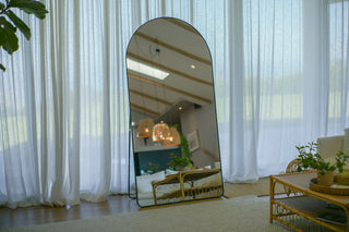 Grandeur Arched Full-Length Mirror (200×100 cm) leaning against a wall, reflecting a bright, modern living space with rattan furniture and soft curtains
