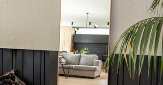 Slim framed full length mirror leaning against a concrete and black timber panelled wall, reflecting a New Zealand living room
