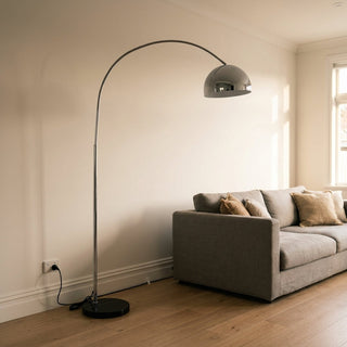 Cosmo chrome arc floor lamp with a black marble base beside a sofa in a New Zealand living room, its cable running along the skirting board to a wall power point