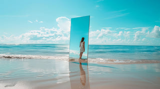 Young woman standing in front of a large mirror on the beach, with a bright blue sky and ocean backdrop, reflecting the summer vibes and vibrant energy of the season