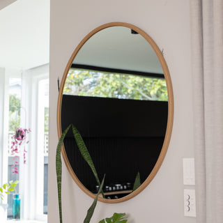 A 100 cm Aure round wall mirror with a natural wooden PS frame and 4 mm double silver glass, mounted on a light grey wall. The mirror reflects a bright, plant-filled modern kitchen, highlighting its clean circular shape and 22 mm slim profile that blends beautifully with contemporary interiors.