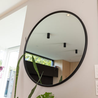 A close-up of the Aure round wall mirror with a sleek black PS frame, highlighting its smooth 22 mm frame depth and 4 mm double silver glass. The mirror reflects a bright modern kitchen and ceiling lights, adding a touch of elegance to the interior space.