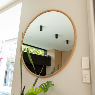 A close-up of the Aure round wall mirror with a warm wooden PS frame, reflecting a bright kitchen and greenery from a nearby window. The mirror’s smooth circular edge and 22 mm depth highlight its modern craftsmanship and subtle elegance, blending perfectly with the light interior decor.