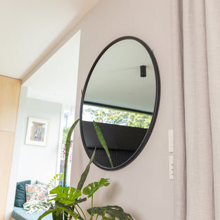 A side view of the Aure round wall mirror with a sleek black PS frame, mounted on a neutral-toned wall beside light curtains. The mirror reflects a bright, airy kitchen with modern decor and a touch of greenery, highlighting its 22 mm frame depth and refined circular design.