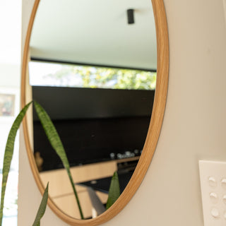 Close-up side angle of the Aure round wall mirror showing its 22 mm deep wooden PS frame and smooth 4 mm double silver glass. The reflection captures a bright modern kitchen with a black splashback, wooden cabinets, and lush indoor greenery adding warmth and elegance to the minimalist space.