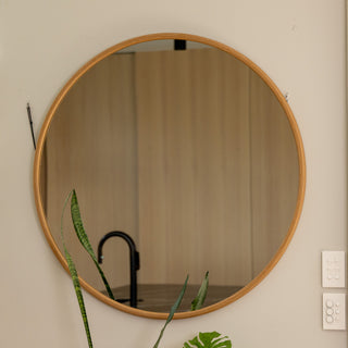 Close-up view of the Aure round wall mirror with a warm wooden PS frame, 22 mm depth, and 4 mm double silver glass. The mirror reflects a modern kitchen interior with soft lighting and a matte black faucet, complemented by indoor plants in the foreground that bring a touch of greenery to the calm, minimalist wall.