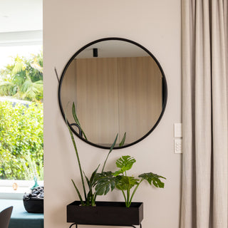 A 100 cm Aure round wall mirror with a modern black PS frame and 22 mm depth, mounted above a black planter filled with lush green indoor plants. The mirror’s 4 mm double silver glass reflects a bright kitchen area and wooden wall panels, adding a touch of style and balance to a clean, natural-themed interior.