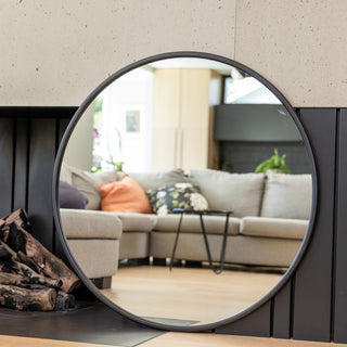 A 100 cm Aure round wall mirror with a sleek black PS frame and 22 mm depth, placed against a modern fireplace wall. The mirror reflects a cosy living room with a grey sofa and colourful cushions, showcasing its 4 mm double silver glass clarity and minimalist elegance in a contemporary home interior.