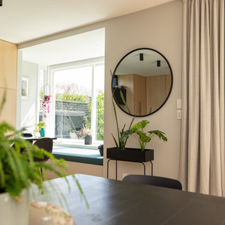 A black-framed Aure round wall mirror, 100 cm in diameter, hanging above a black planter in a bright modern dining room. The 22 mm deep PS frame and 4 mm double silver glass reflect the airy window light and greenery, blending sleek design with natural warmth for a refined interior look.