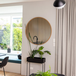 A 100 cm Aure round wall mirror with a natural wooden PS frame, hanging above a black planter in a bright modern dining space. The mirror reflects the soft light from nearby windows and a sleek kitchen area, showcasing its slim 22 mm frame depth and smooth 4 mm double silver glass finish that adds warmth and clarity to the room.