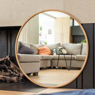 A 100 cm Aure round wall mirror with a warm wooden PS frame, 22 mm deep, and 4 mm double silver glass. The mirror reflects a cosy modern living room with a beige sofa, soft cushions, and natural light, showcasing the mirror’s clarity and timeless circular design.