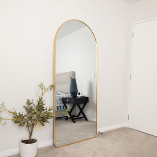 Front view of the Aurelia arched full-length mirror with slim gold frame, reflecting a modern bedroom with bedside table and décor.