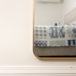 Close-up of the Aurelia arched full-length mirror showing its smooth rounded gold frame corner against a white wall, reflecting part of a bedroom setting.