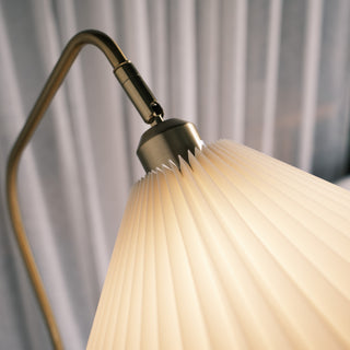 Close up of the Aurelia floor lamp’s pleated fabric shade, showing the warm diffused light, brushed brass fitting, and curved metal arm against soft grey curtains.
