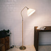 Aurelia Pleated Fabric Floor Lamp featuring a soft cream pleated shade, a curved brass arm, and a wood-print metal pole with a brass base. The lamp is shown standing against a white brick wall beside a wooden desk and a side table with a dark planter, creating a warm, minimalist interior setting.