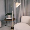 Aurelia Pleated Fabric Floor Lamp featuring a pleated cream fabric shade and curved brass neck, paired with a wood-print metal pole and brass base. The lamp stands beside a round wooden side table with stacked books and a potted plant, set in a soft neutral living room with curtains and a light upholstery sofa.