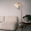 Aurelia Pleated Fabric Floor Lamp with a cream pleated shade, curved brass arm, wood textured pole, and brass base. The lamp is placed beside a soft beige sofa and a round wooden side table with books and a potted plant, set against a white painted brick wall in a warm, modern living room.