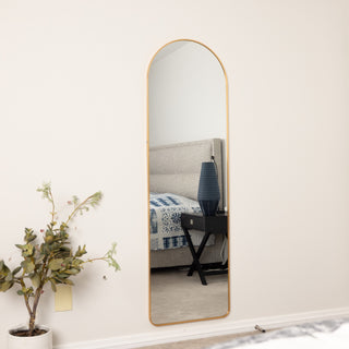 The smaller 160x50cm Aurelia arched full-length mirror with a gold frame, styled against a white wall beside a plant, reflecting a modern bedroom interior with a bed and blue lamp.