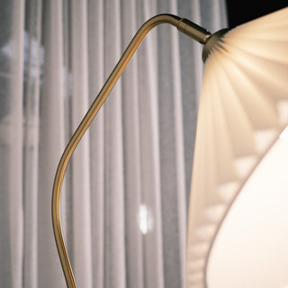 Close up of the Aurelia floor lamp’s curved brass metal arm beside the pleated fabric shade, with soft curtains in the background creating a warm and elegant look.
