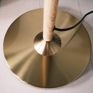 Close up of the Aurelia Pleated Fabric Floor Lamp base, showing the brushed brass metal plate and the wood textured pole connection, with the black cord visible against a light floor surface.