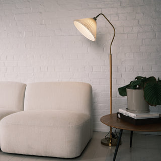 Aurelia Pleated Fabric Floor Lamp with a cream pleated shade, curved brass arm, wood textured pole, and brass base. The lamp is placed beside a soft beige sofa and a round wooden side table with books and a potted plant, set against a white painted brick wall in a warm, modern living room.