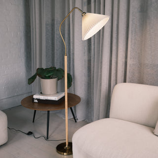Aurelia Pleated Fabric Floor Lamp featuring a pleated cream fabric shade and curved brass neck, paired with a wood-print metal pole and brass base. The lamp stands beside a round wooden side table with stacked books and a potted plant, set in a soft neutral living room with curtains and a light upholstery sofa.