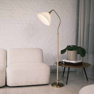 Aurelia Pleated Fabric Floor Lamp featuring a cream pleated shade, curved brass arm, wood textured pole, and brass base. The lamp sits beside a soft beige sofa and a round wooden side table with books and a potted plant, against a white painted brick wall with soft curtains in a calm, modern living room.

