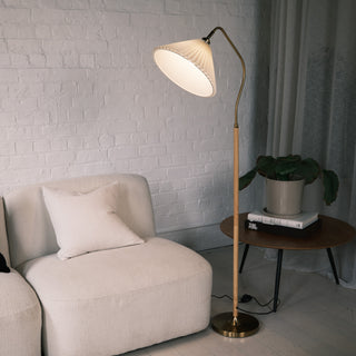 Aurelia Pleated Fabric Floor Lamp featuring a soft pleated fabric shade, curved warm brass neck, and wood-print metal pole with a brass base. The lamp is placed beside a cream lounge chair and a round wooden side table with a plant, set against a white brick wall and soft curtains in a modern New Zealand living room.
