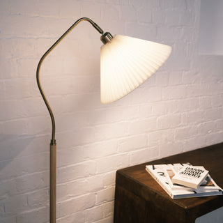The Aurelia floor lamp with a pleated fabric shade casting a warm light, featuring a brushed brass curved arm and wood textured pole beside a wooden console with stacked books in a soft interior setting.