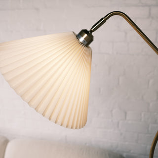 Close up of the Aurelia floor lamp’s pleated cream fabric shade, showing the brushed brass fitting and warm diffused light against a soft white brick wall.