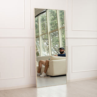 The Branewood 180x80cm full-length mirror featuring a natural textured PS wood-look frame and 4mm clear glass, placed against a white panelled wall reflecting a bright modern living room with large windows. Designed for both wall mounting and floor use, this mirror brings a warm, minimalist charm to contemporary New Zealand interiors.