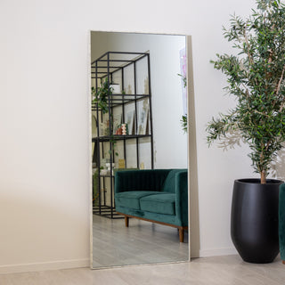 Branewood 180x80cm full-length mirror with a rustic light wood-textured PS frame reflecting a modern living room with a green velvet sofa, open shelving, and indoor plants. The 4mm clear glass provides crisp reflection, ideal for both wall-mounted and floor styling in elegant New Zealand homes.